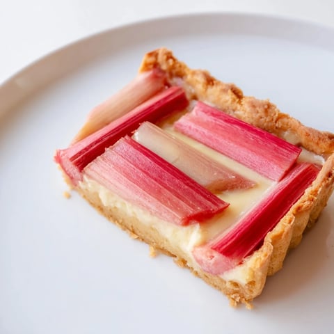 A close-up of the Rhubarb, White Chocolate, and Elderflower Tart, featuring bright pink roasted rhubarb slices arranged over a creamy, pale yellow custard in a golden-brown tart shell.